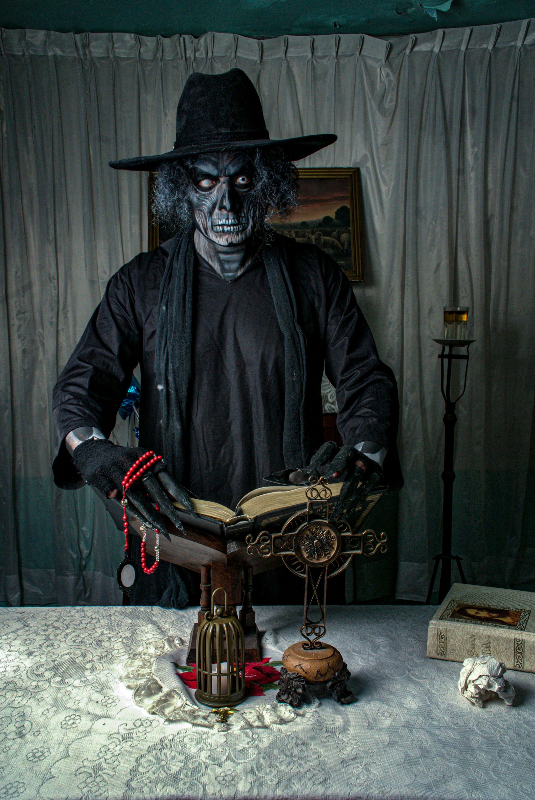 A costumed figure with a skeletal face reads a ritual book surrounded by gothic decor indoors.